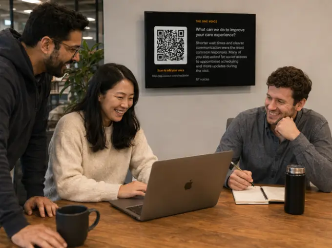 Three people around a laptop at a wooden table, laughing, with a wall display behind them showing a voicer's QR code and one voice