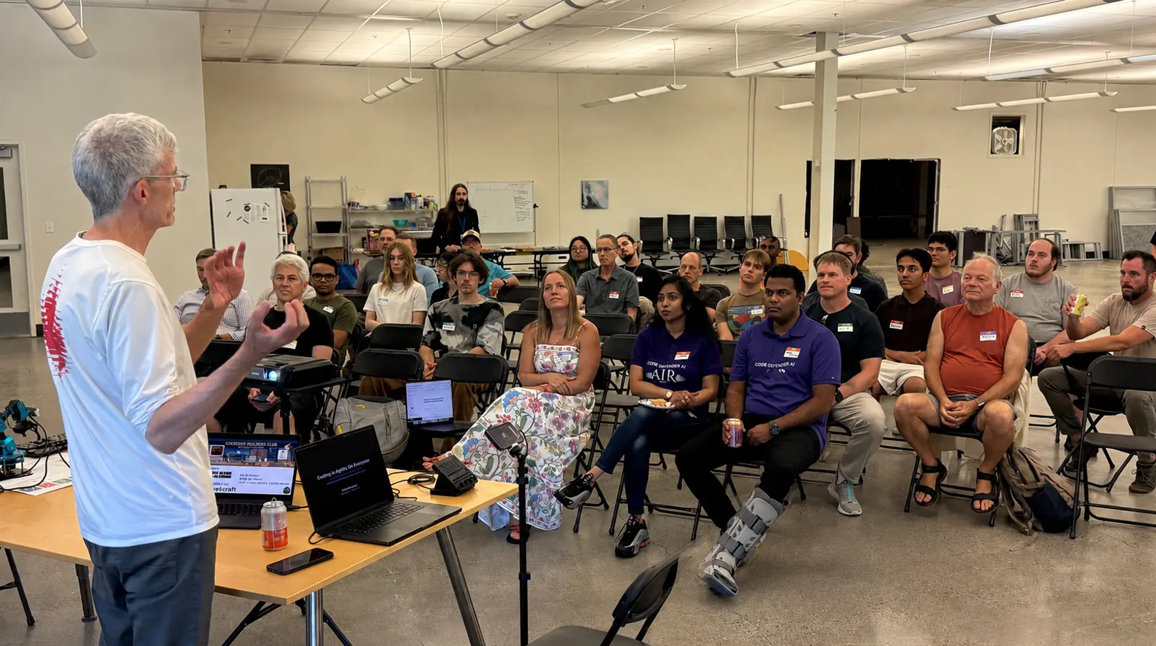 Sam Keen stands at a laptop table and speaks to about twenty-five builders seated on folding chairs in a large open room, with a small robot arm on the demo table beside him.