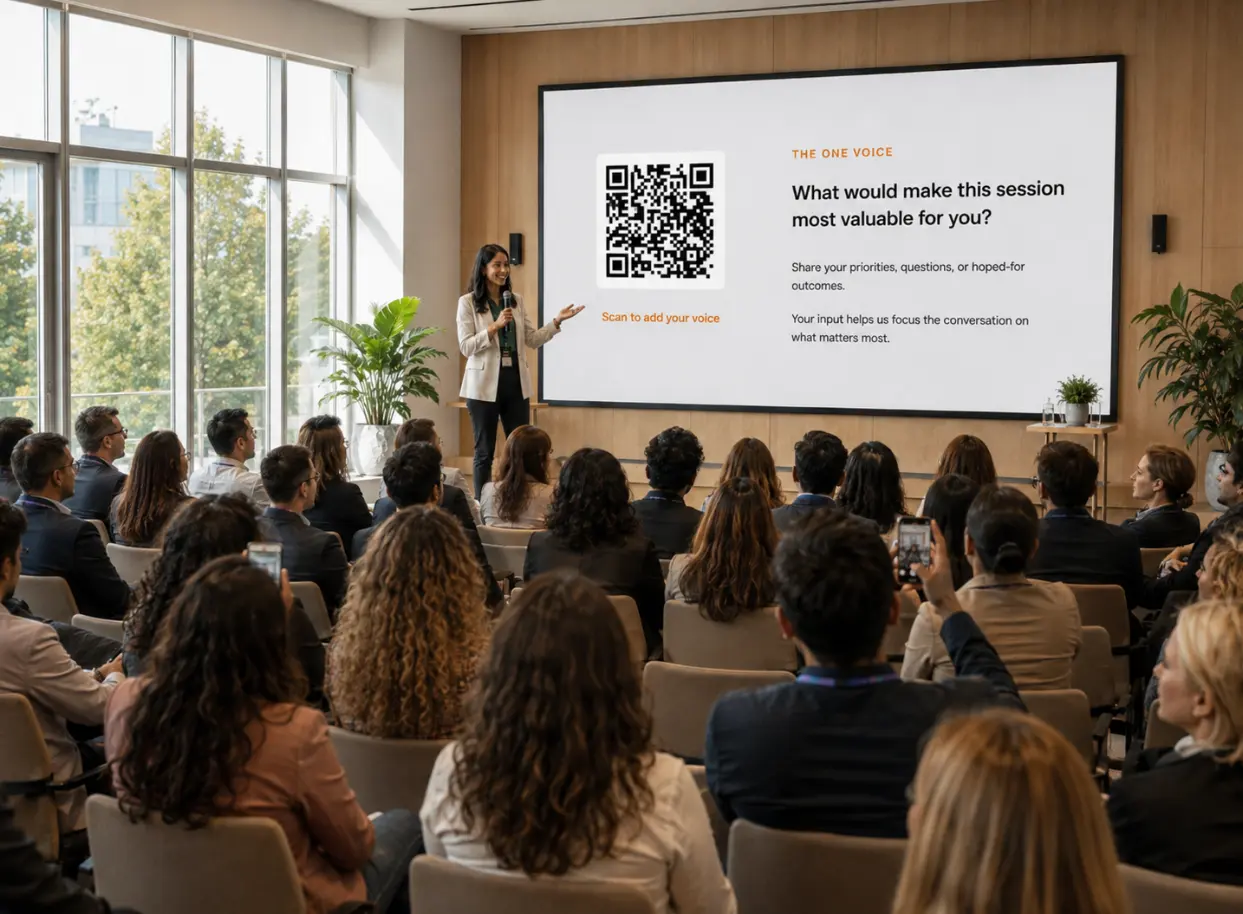 A facilitator on stage in front of a large seated audience, presenting a One Voicer screen with a QR code and the question “What would make this session most valuable for you?”