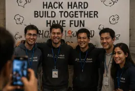 Five smiling hackathon teammates in matching shirts and lanyards posing for a phone photo in front of a banner reading “Hack hard, build together, have fun.”