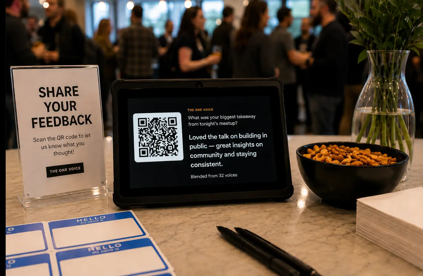 A meetup welcome table with name tags, snacks, a sign reading “Share your feedback: scan the QR code to let us know what you thought!”, and a tablet showing a question, a QR code, and an answer blended from 32 voices.