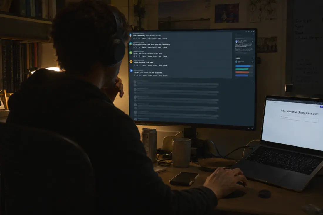 A moderator at a desk at night, headphones on, facing a monitor where a heated thread shows five active comments above a long faded scroll of empty rows, with a laptop beside it showing the question “What should we change this month?”