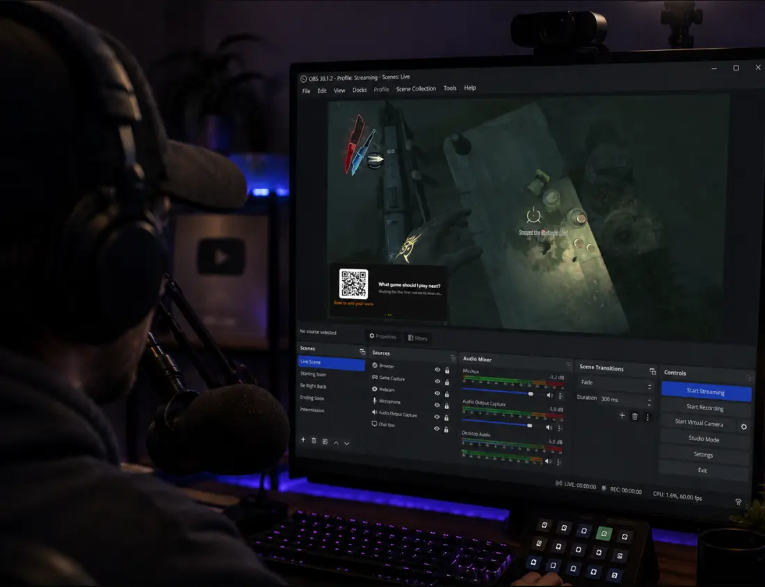 A streamer at a desk wearing headphones, with OBS open on the monitor and a small overlay in the corner of the game preview showing a QR code and the question “What game should I play next?”