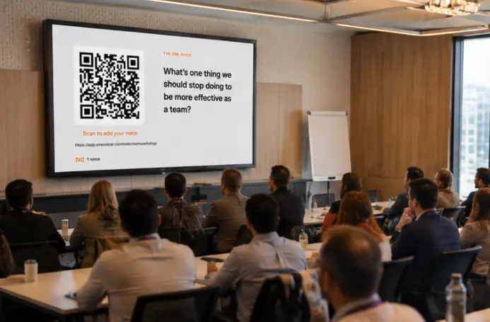 A team in a conference room watching a large screen showing a One Voicer question, “What’s one thing we should stop doing to be more effective as a team?”, with a scan-to-answer QR code beside it.
